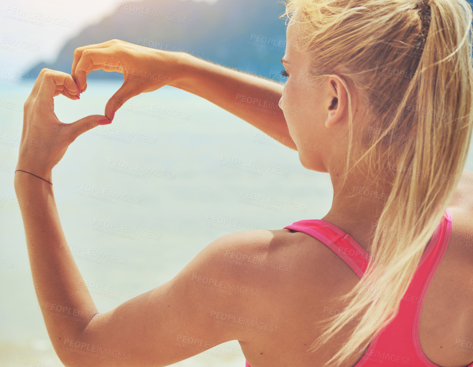 Buy stock photo Rearview shot of a sporty young woman making a heart shape with her hands while standing on the beach