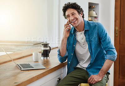 Buy stock photo Shot of a man speaking on his cellphone while using his laptop in the kitchen