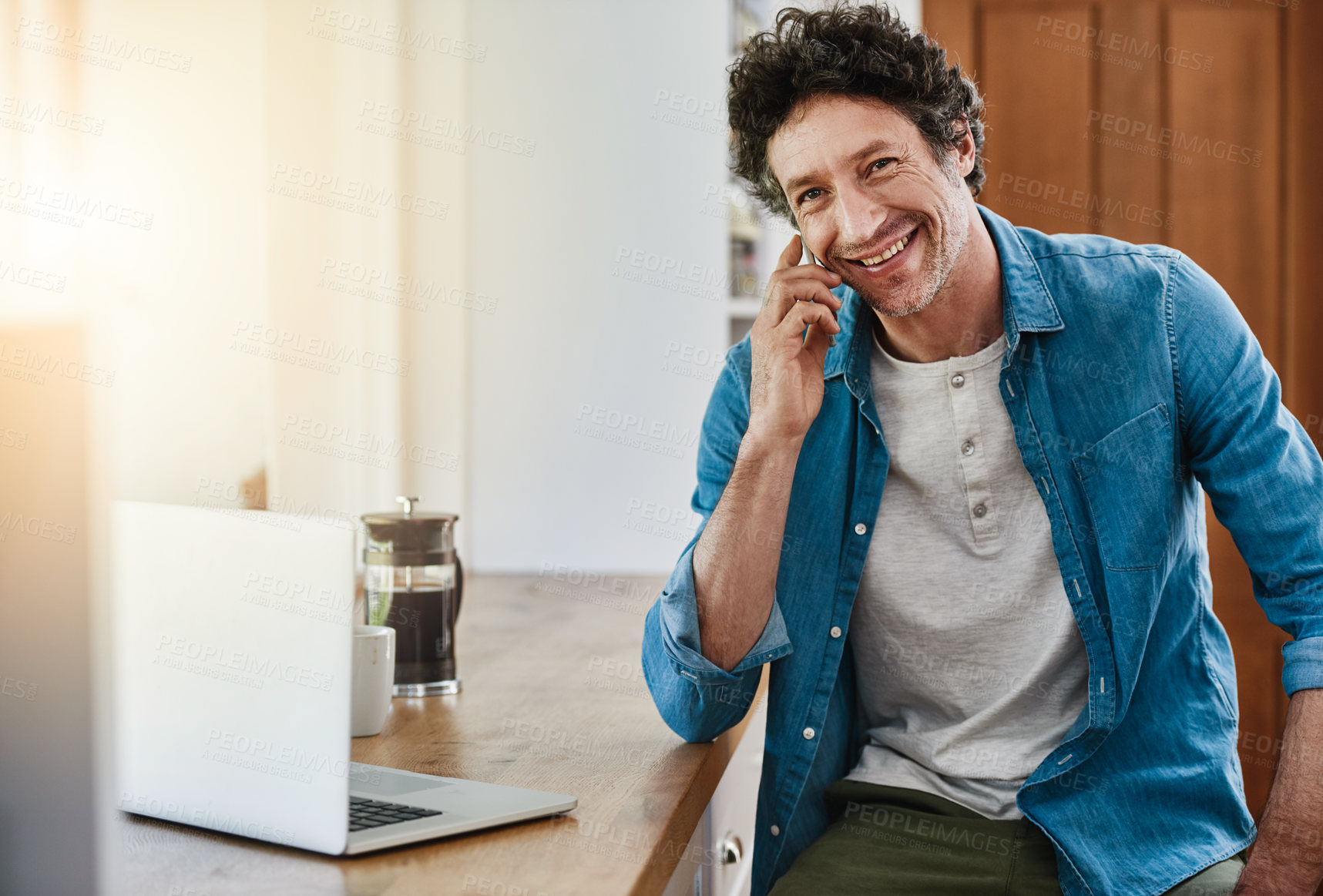 Buy stock photo Portrait of a man speaking on his cellphone while using his laptop in the kitchen