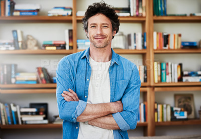 Buy stock photo Shot of a happy bachelor posing with his arms crossed in front of a bookshelf at home