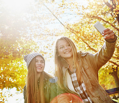 Buy stock photo Shot of two young friends taking selfies together on a mobile phone while out in the park