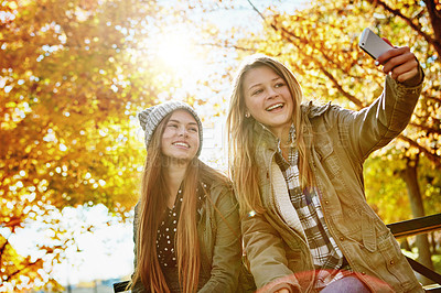 Buy stock photo Shot of two young friends taking selfies together on a mobile phone while out in the park