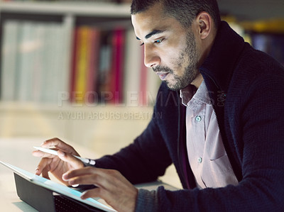 Buy stock photo Shot of a focused young man working on a digital tablet in his home office in the early evening
