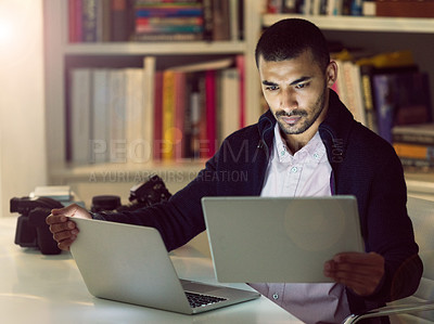 Buy stock photo Shot of a focused young photographer working on a laptop and digital tablet in his home office in the early evening