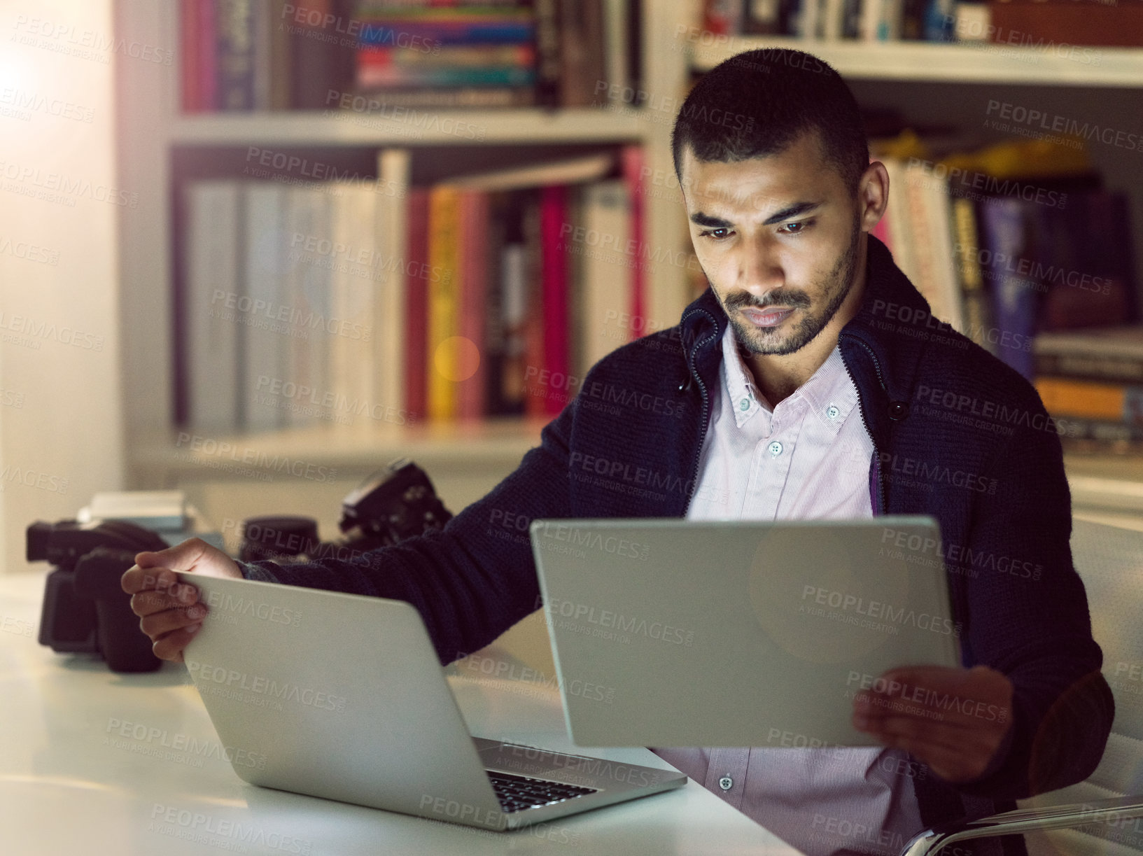 Buy stock photo Shot of a focused young photographer working on a laptop and digital tablet in his home office in the early evening