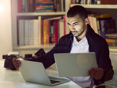 Buy stock photo Shot of a focused young photographer working on a laptop and digital tablet in his home office in the early evening