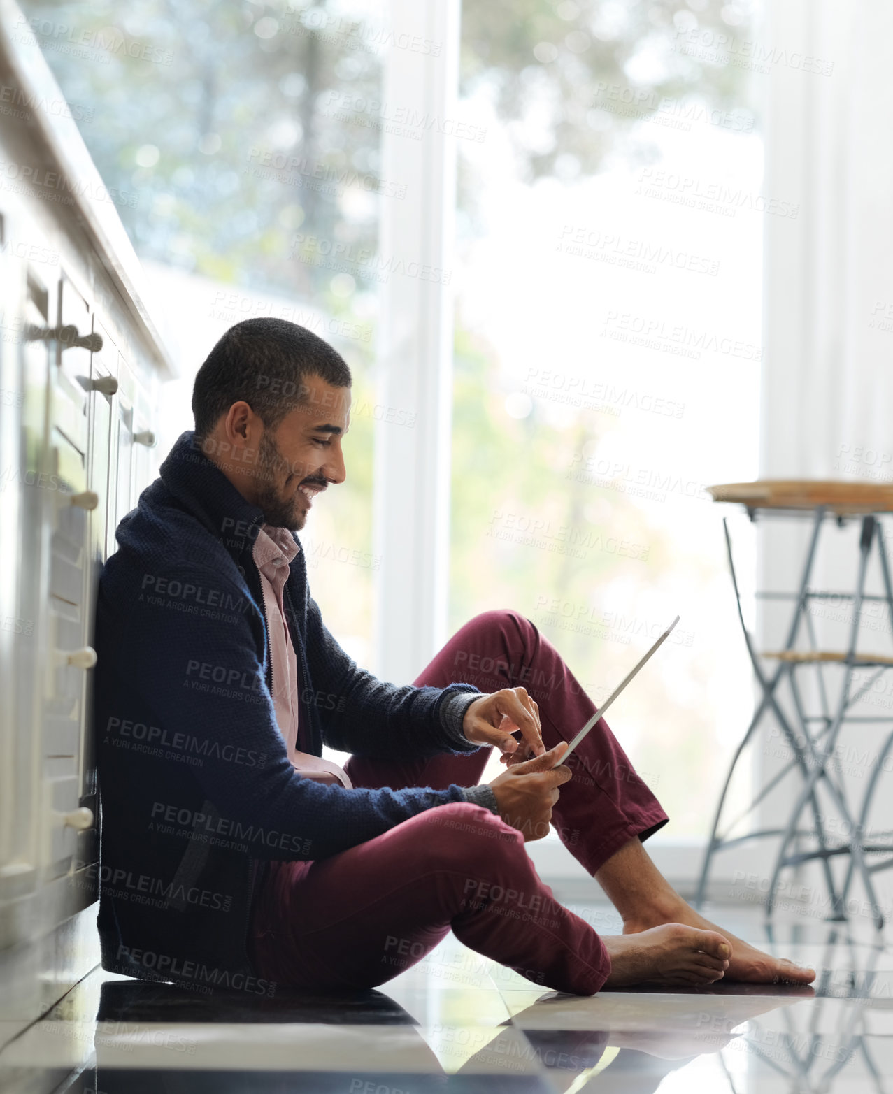 Buy stock photo Shot of a smiling young man sitting on his kitchen floor using a digital tablet