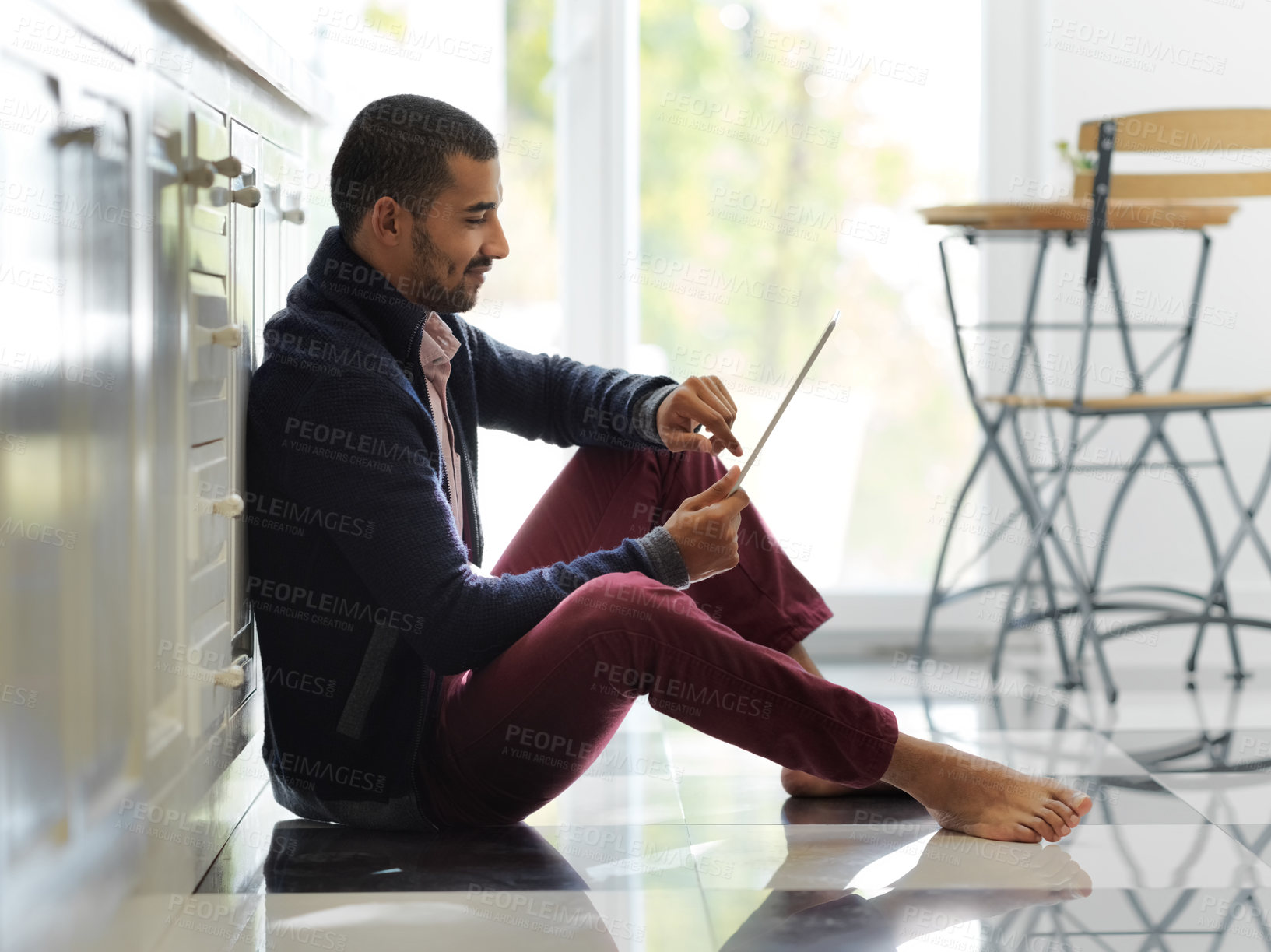 Buy stock photo Shot of a smiling young man sitting on his kitchen floor using a digital tablet