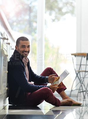 Buy stock photo Portrait of a smiling young man sitting on his kitchen floor using a digital tablet