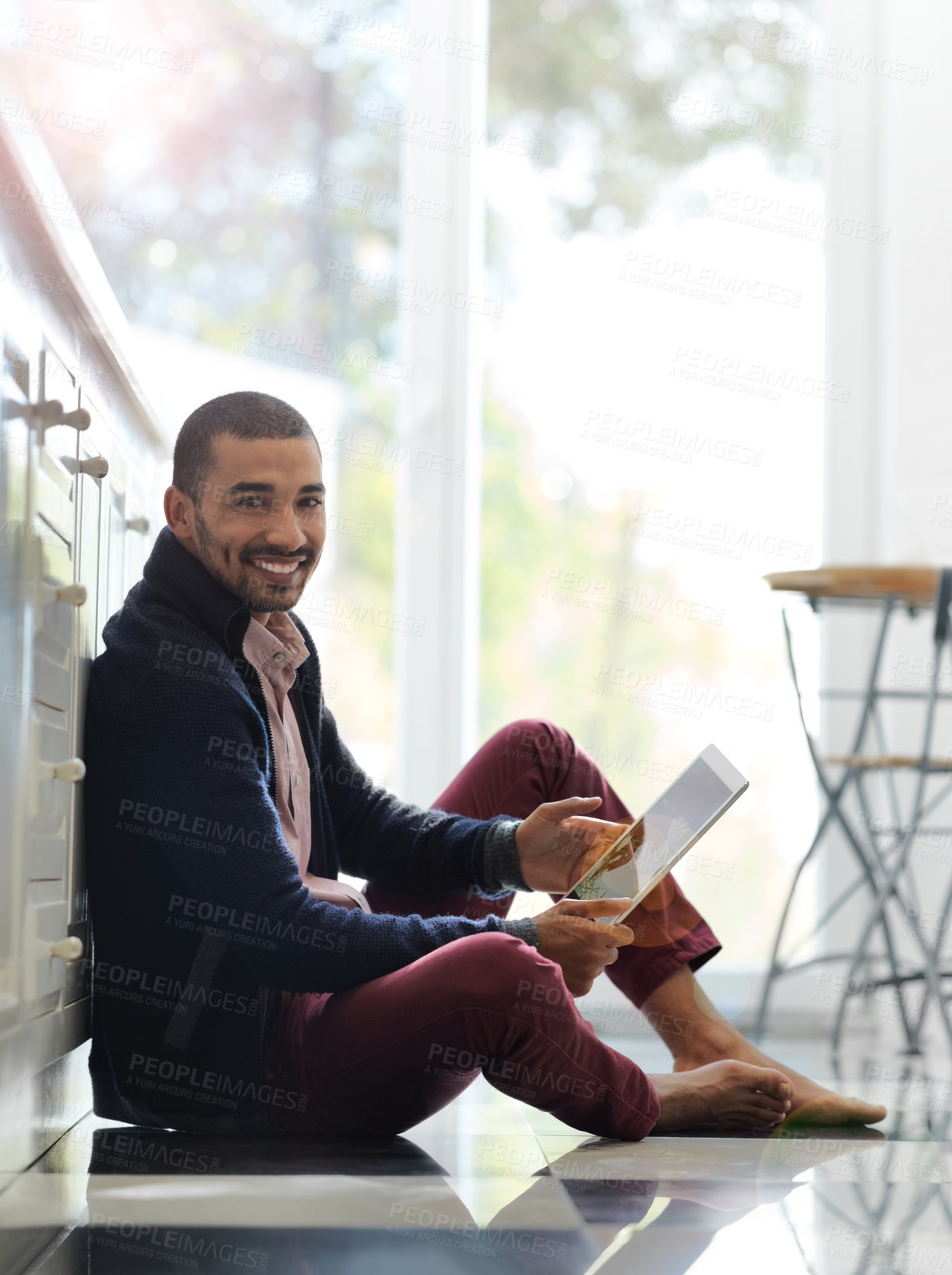 Buy stock photo Portrait of a smiling young man sitting on his kitchen floor using a digital tablet