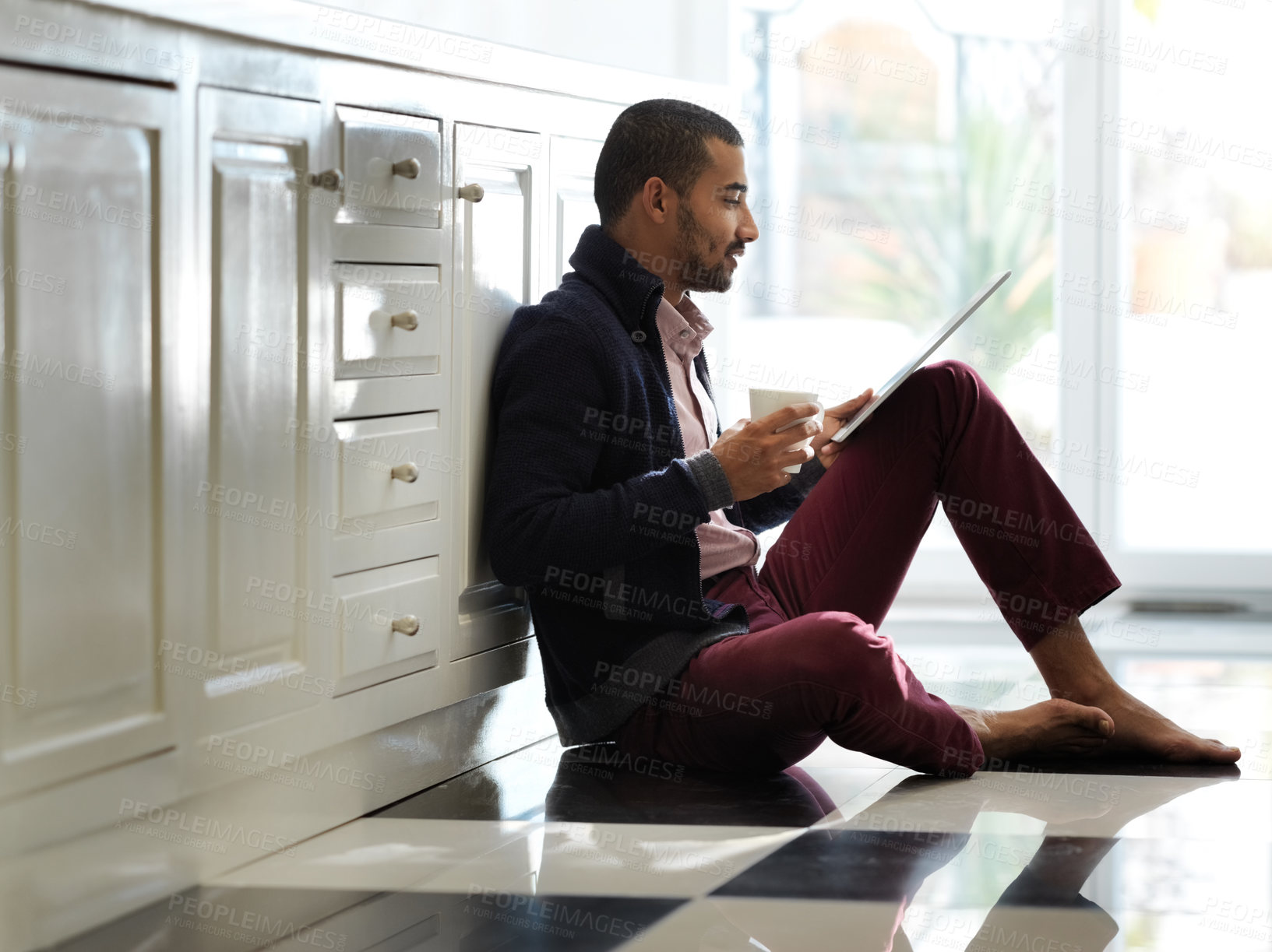 Buy stock photo Shot of a smiling young man sitting on his kitchen floor using a digital tablet