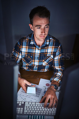 Buy stock photo High angle shot of a young programmer holding his smartphone while using his computer in the office
