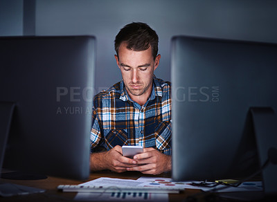 Buy stock photo Shot of a young programmer using his smartphone while sitting behind his computer in the office