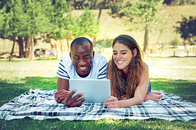 Buy stock photo Interracial couple, relax and blanket with tablet on grass field for outdoor browsing, entertainment or social media. Young man and woman with smile and lying on picnic mat with technology in nature