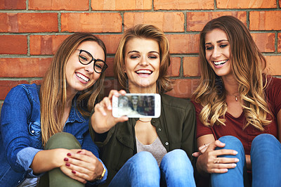 Buy stock photo Shot of three smiling female university students taking a selfie together while sitting outside on campus