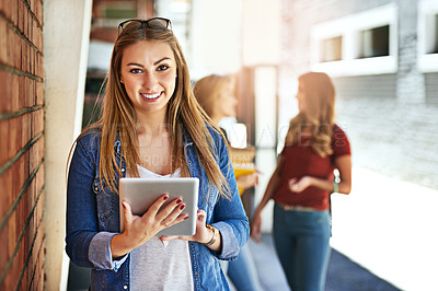 Buy stock photo Shot of a smiling female university student standing on campus using a digital tablet with friends in the background
