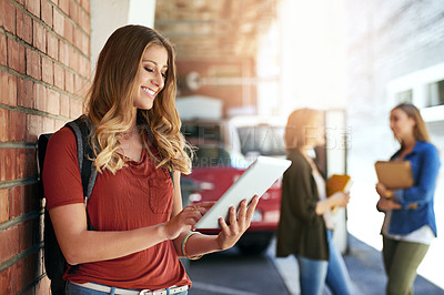 Buy stock photo Shot of a smiling female university student standing on campus using a digital tablet with friends in the background