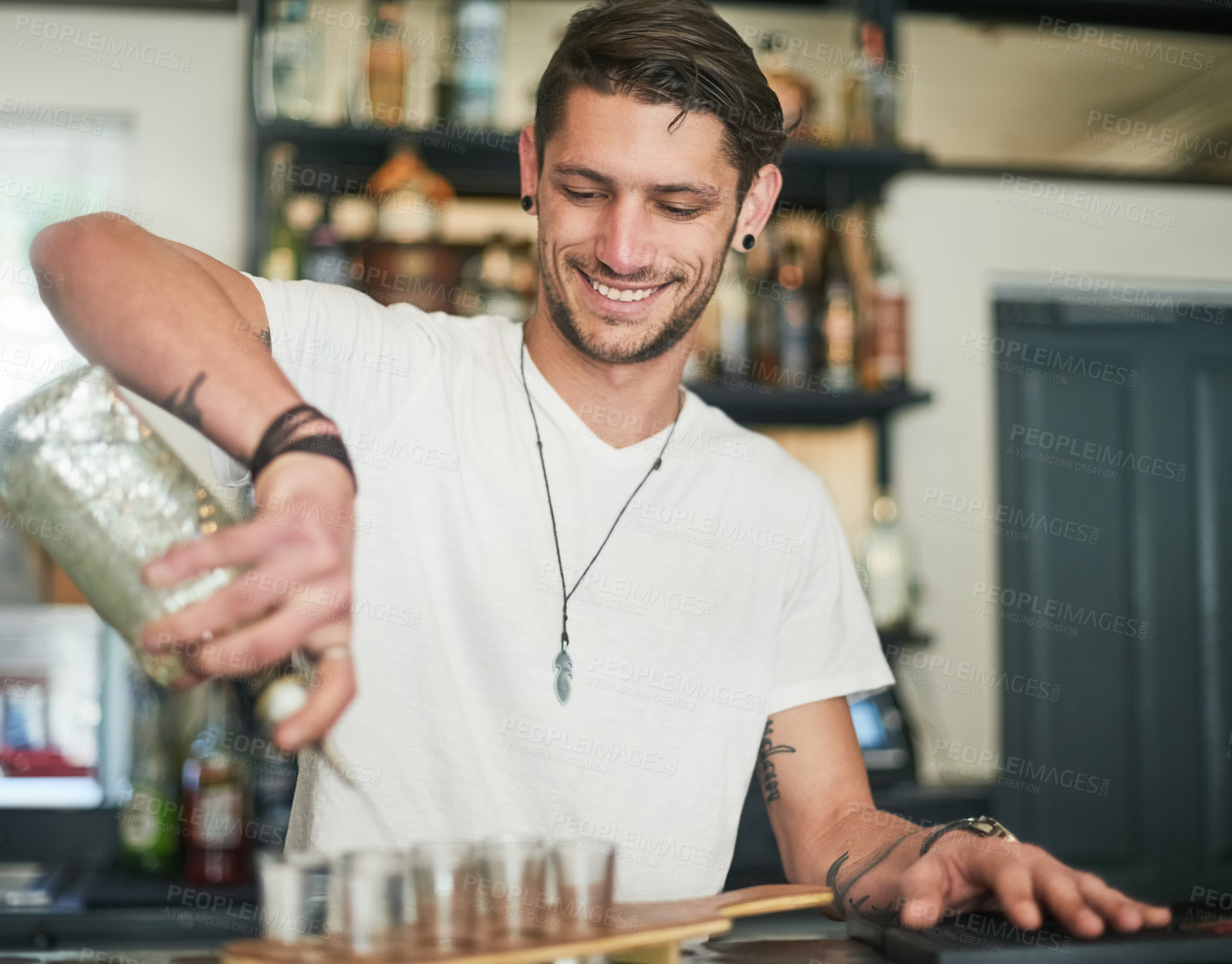 Buy stock photo Shot of a happy young bartender pouring shots behind the bar