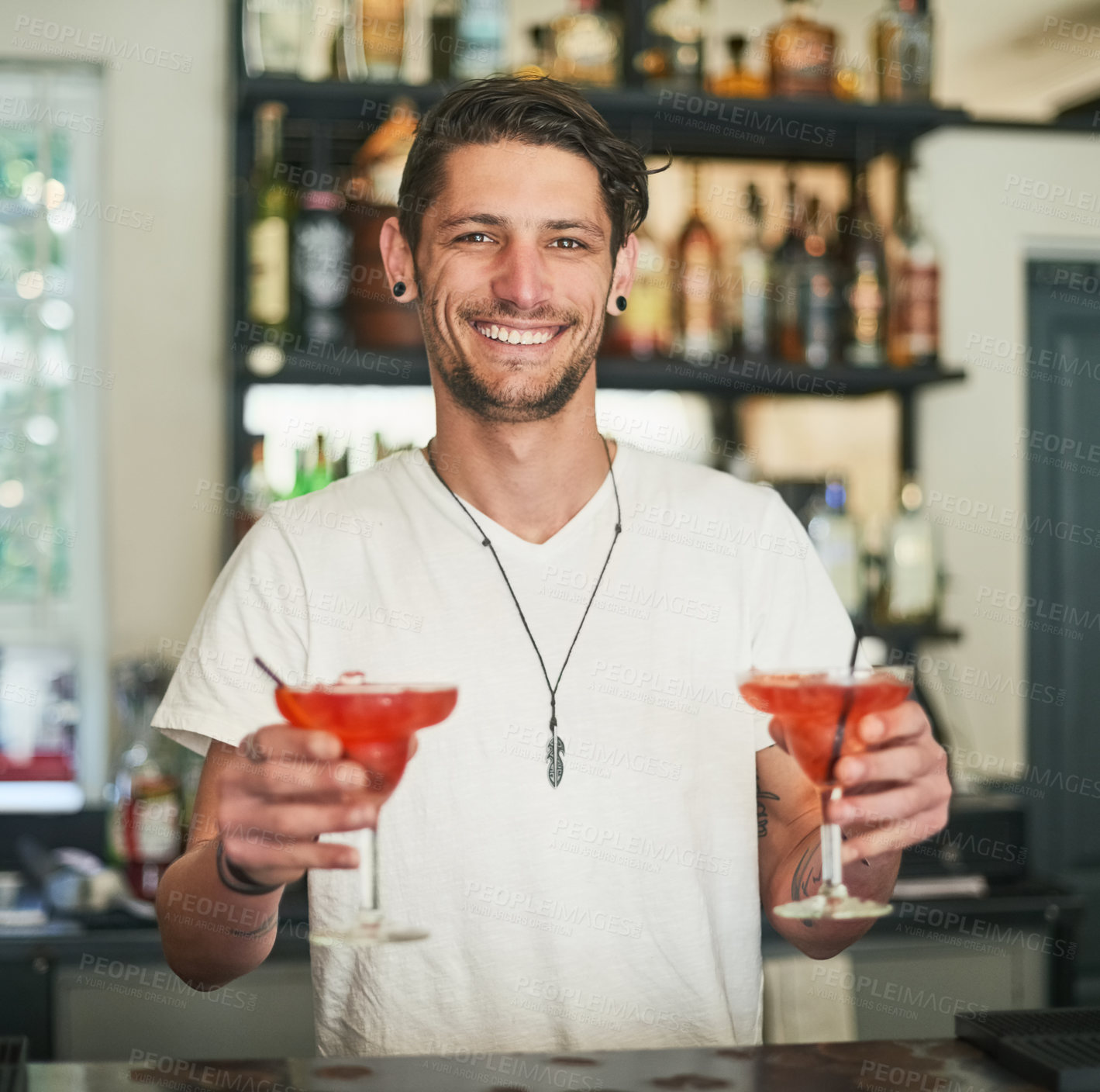 Buy stock photo Portrait of a happy young bartender holding two cocktails while standing behind the bar