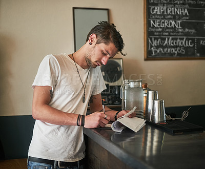 Buy stock photo Shot of a focussed young bartender making notes on receipts behind the bar
