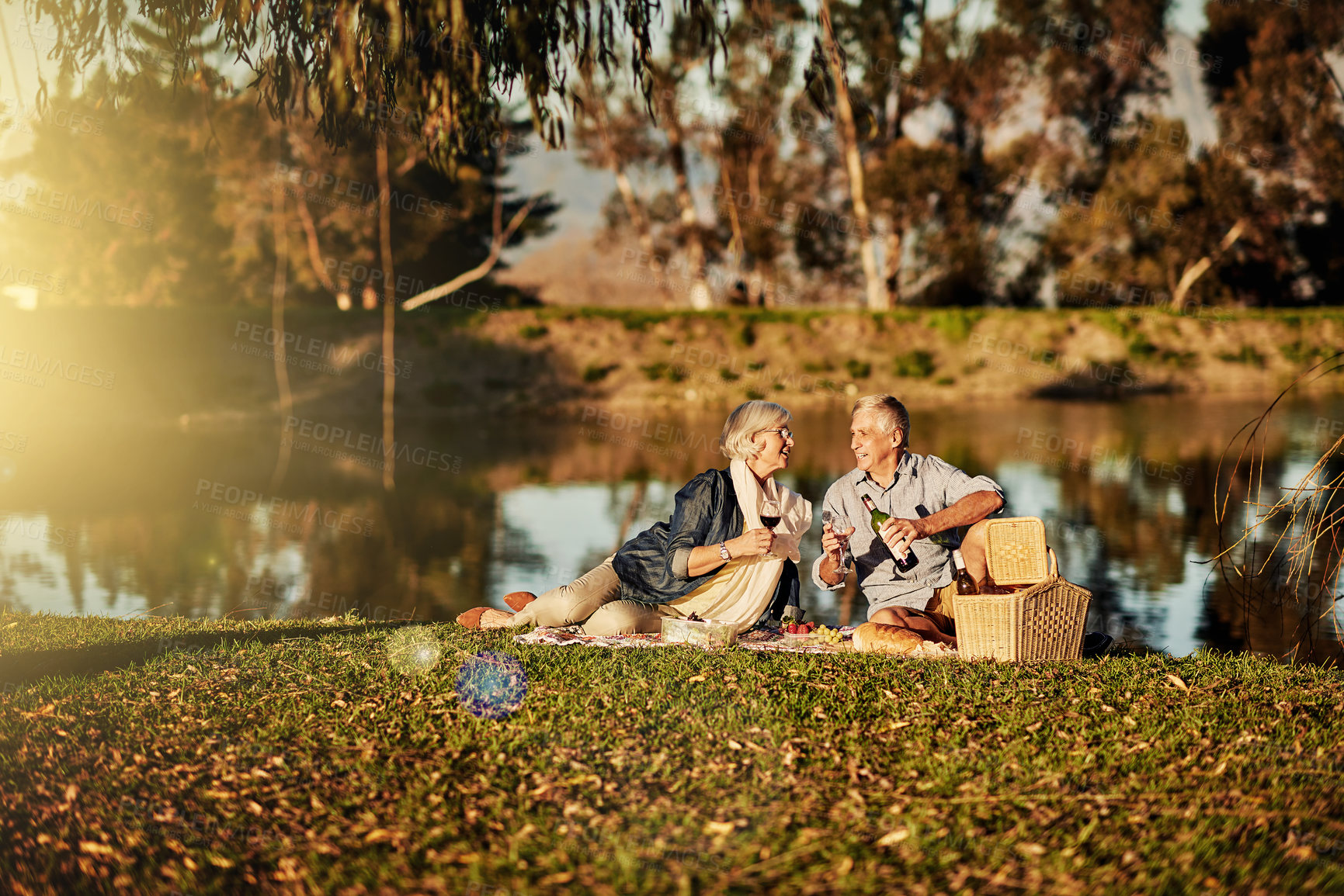 Buy stock photo Senior couple, alcohol and picnic in outdoor for love, romance and relax by lake in nature. Elderly people, speaking and drink wine on vacation, holiday and calm retirement for bonding by river