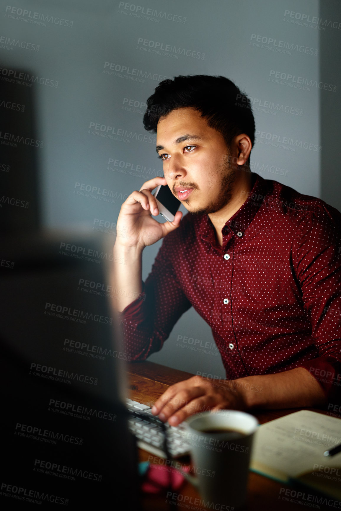 Buy stock photo Shot of a young male designer talking on the phone and working on his computer late at night
