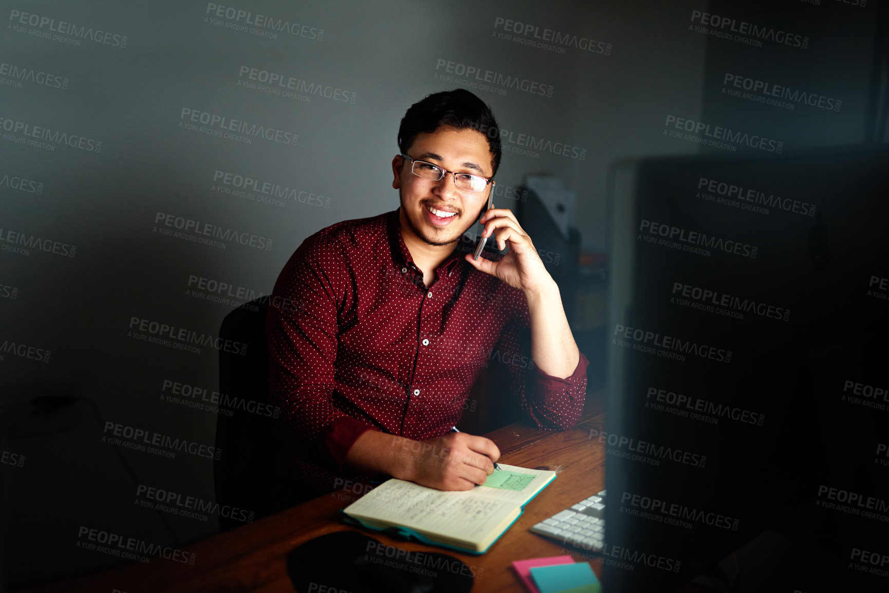 Buy stock photo Portrait of a young male designer talking on the phone and working on his computer late at night