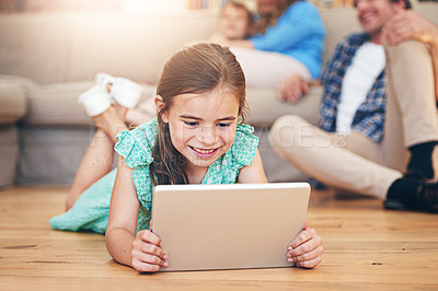 Buy stock photo Shot of a little girl lying on the floor and using a digital tablet with her family in the background