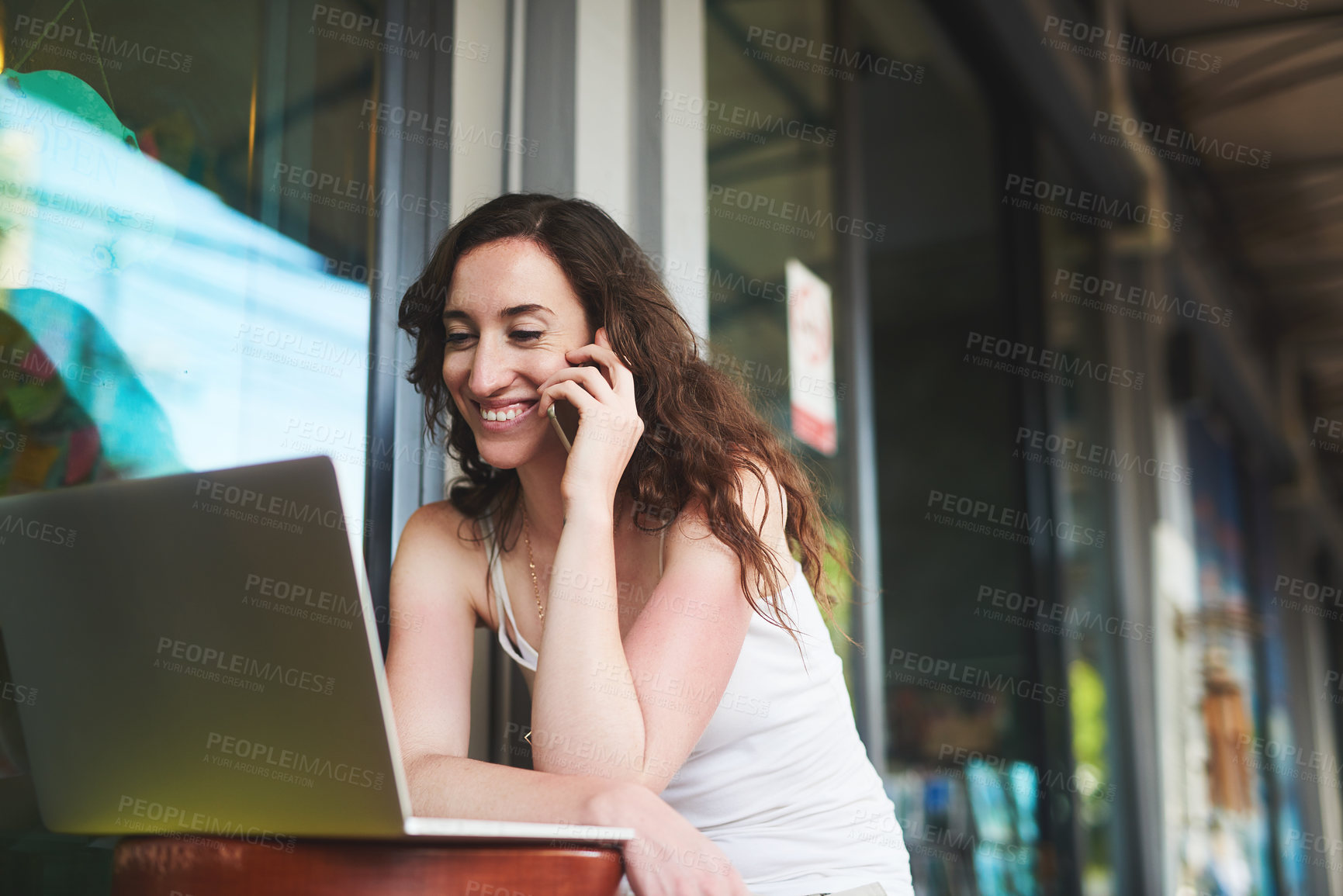Buy stock photo Shot of a young woman speaking on her cellphone while using her laptop outside a coffee shop