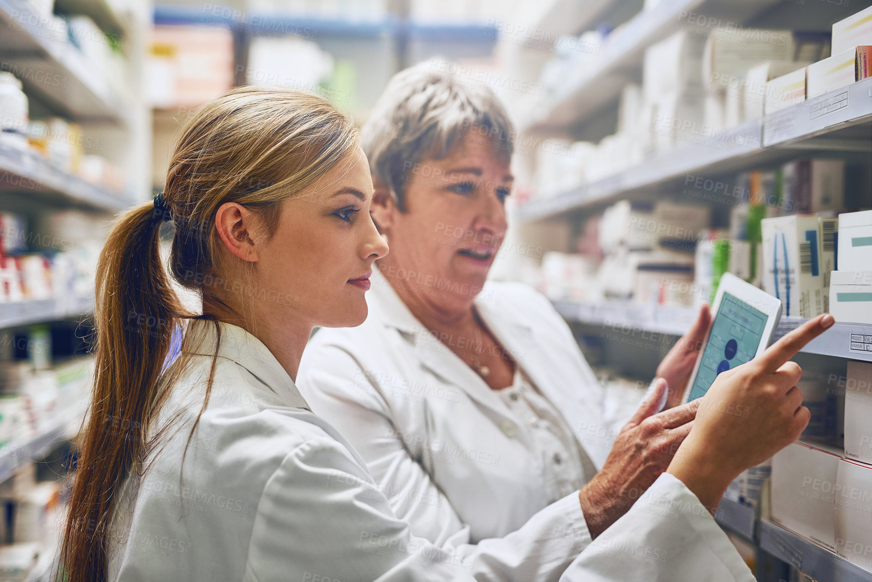 Buy stock photo Shot of two pharmacists using a digital tablet while checking stock on the shelf