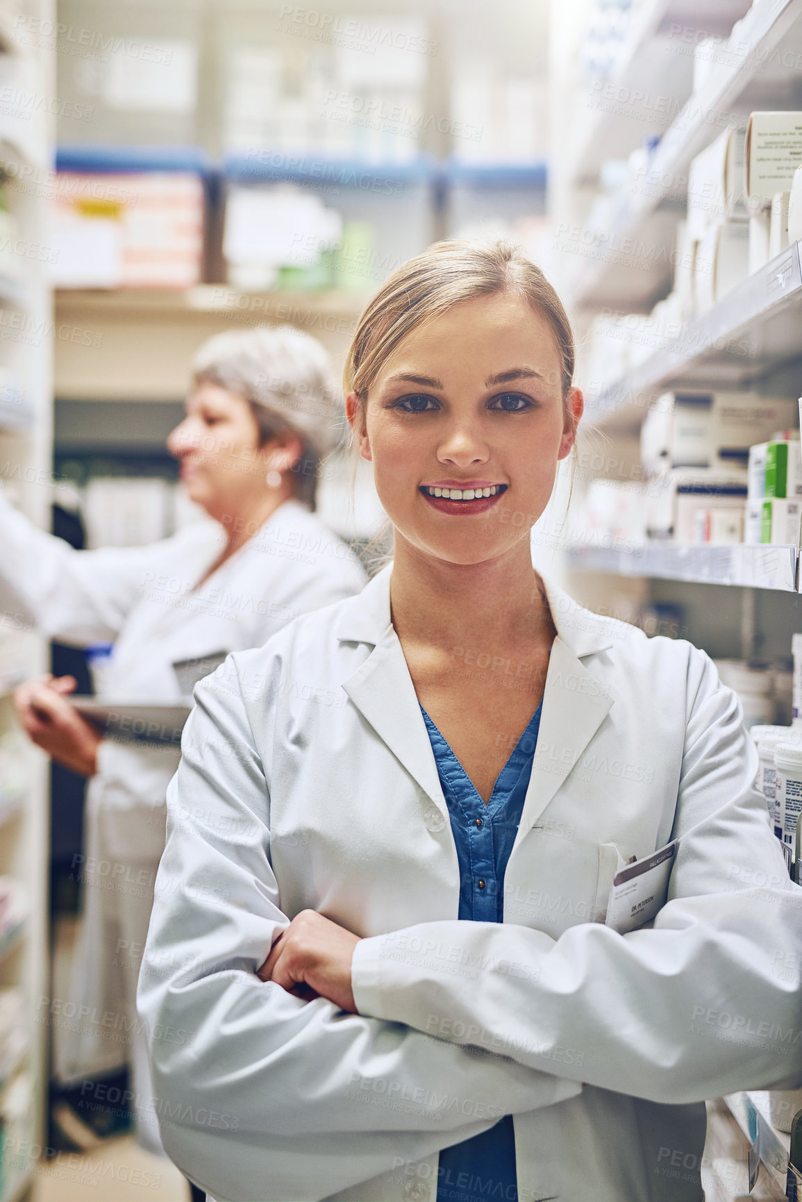 Buy stock photo Portrait of a pharmacist standing in a isle with her colleague working in the background