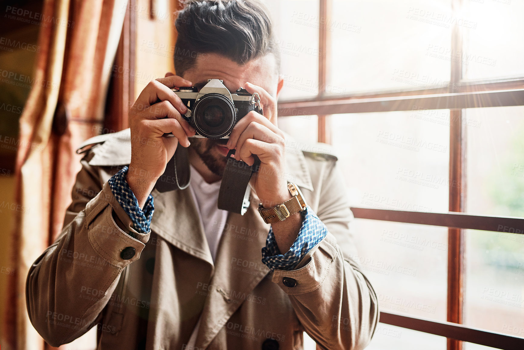 Buy stock photo Shot of a handsome young man  standing by a window at home using a camera