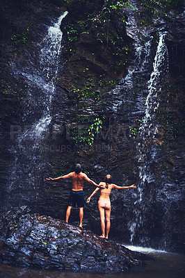 Buy stock photo Rearview shot of a young couple standing under a waterfall in the jungle