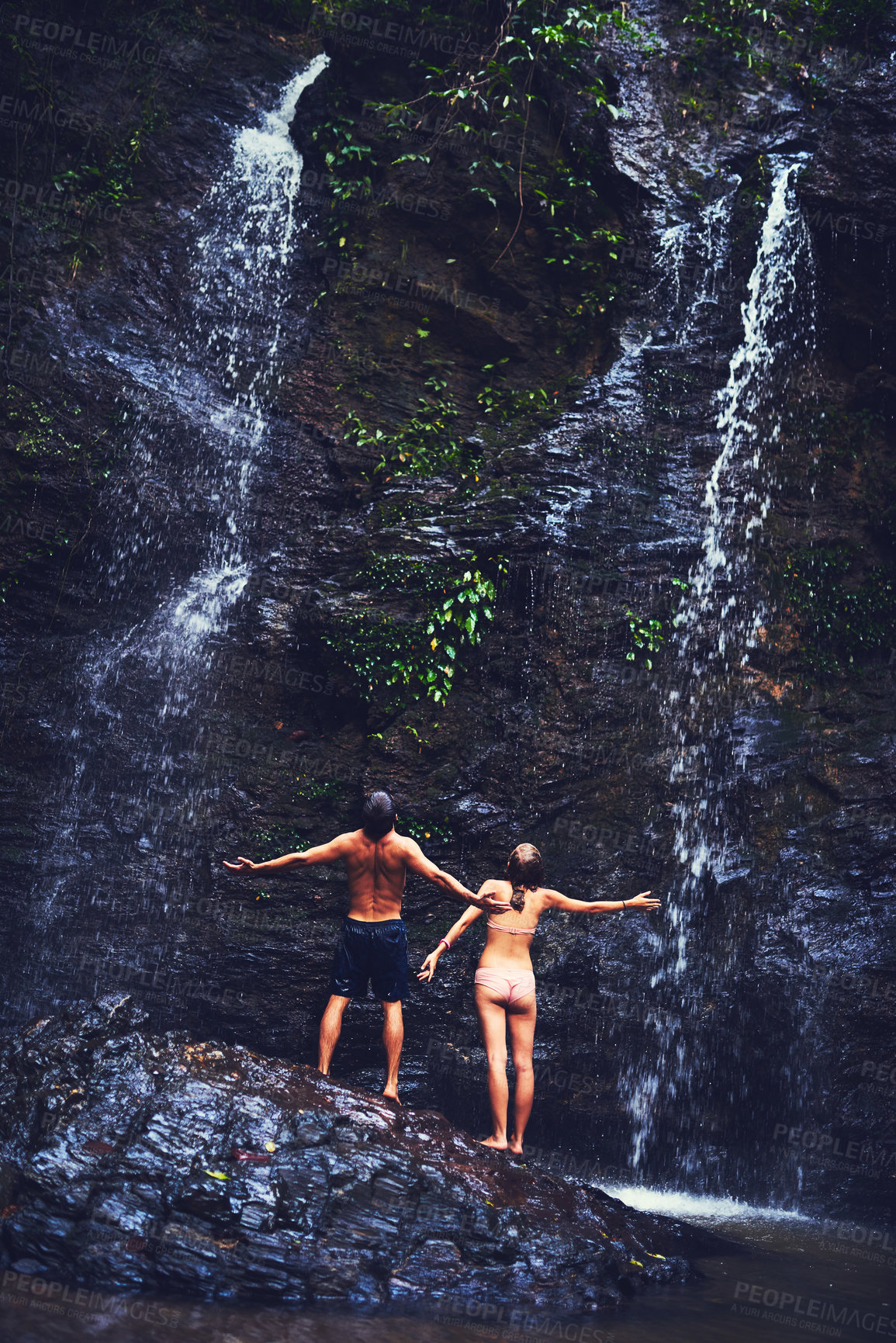 Buy stock photo Rearview shot of a young couple standing under a waterfall in the jungle
