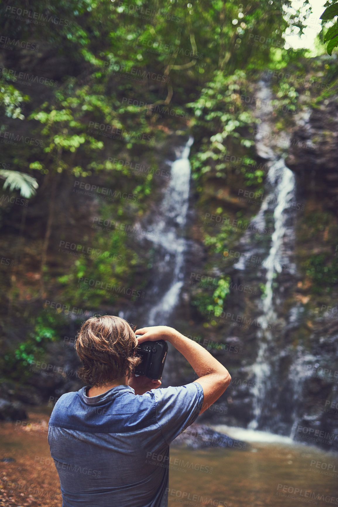 Buy stock photo Rearview shot of a young photographer taking a picture of a waterfall in the jungle