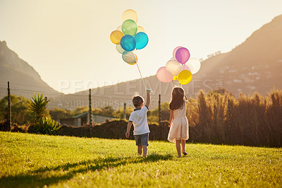 Buy stock photo Rearview shot of two cute little siblings holding balloons while walking outside