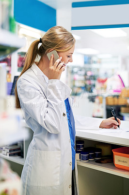 Buy stock photo Shot of a young pharmacist talking on a cellphone while filling out a prescription in a chemist