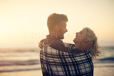 Buy stock photo Couple, hug and laughing at beach, sunset and happy for connection, joke and relax with blanket, Man, woman and partner with embrace for love, care and funny memory on vacation by sea in sunshine