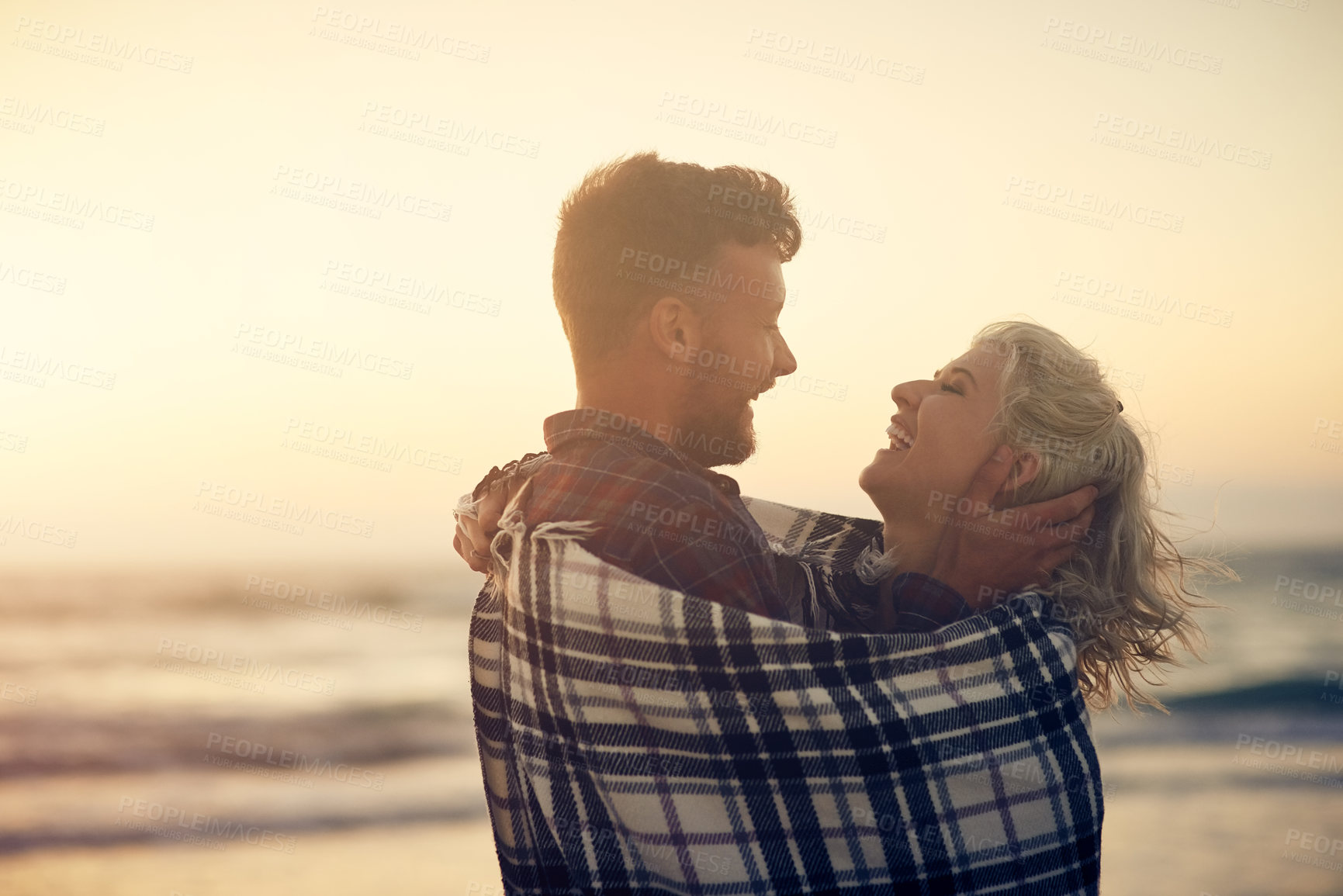 Buy stock photo Couple, hug and laughing at beach, sunset and happy for connection, joke and relax with blanket, Man, woman and partner with embrace for love, care and funny memory on vacation by sea in sunshine