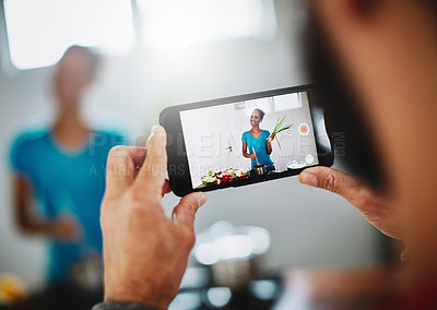 Buy stock photo Shot of a man filming his wife on his smartphone while she cooks in their kitchen at home