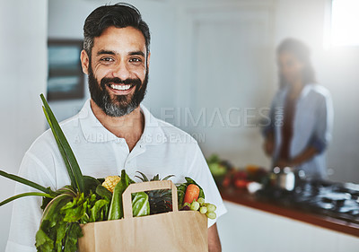 Buy stock photo Portrait of a happy man holding a bag of fresh vegetables while standing in his kitchen