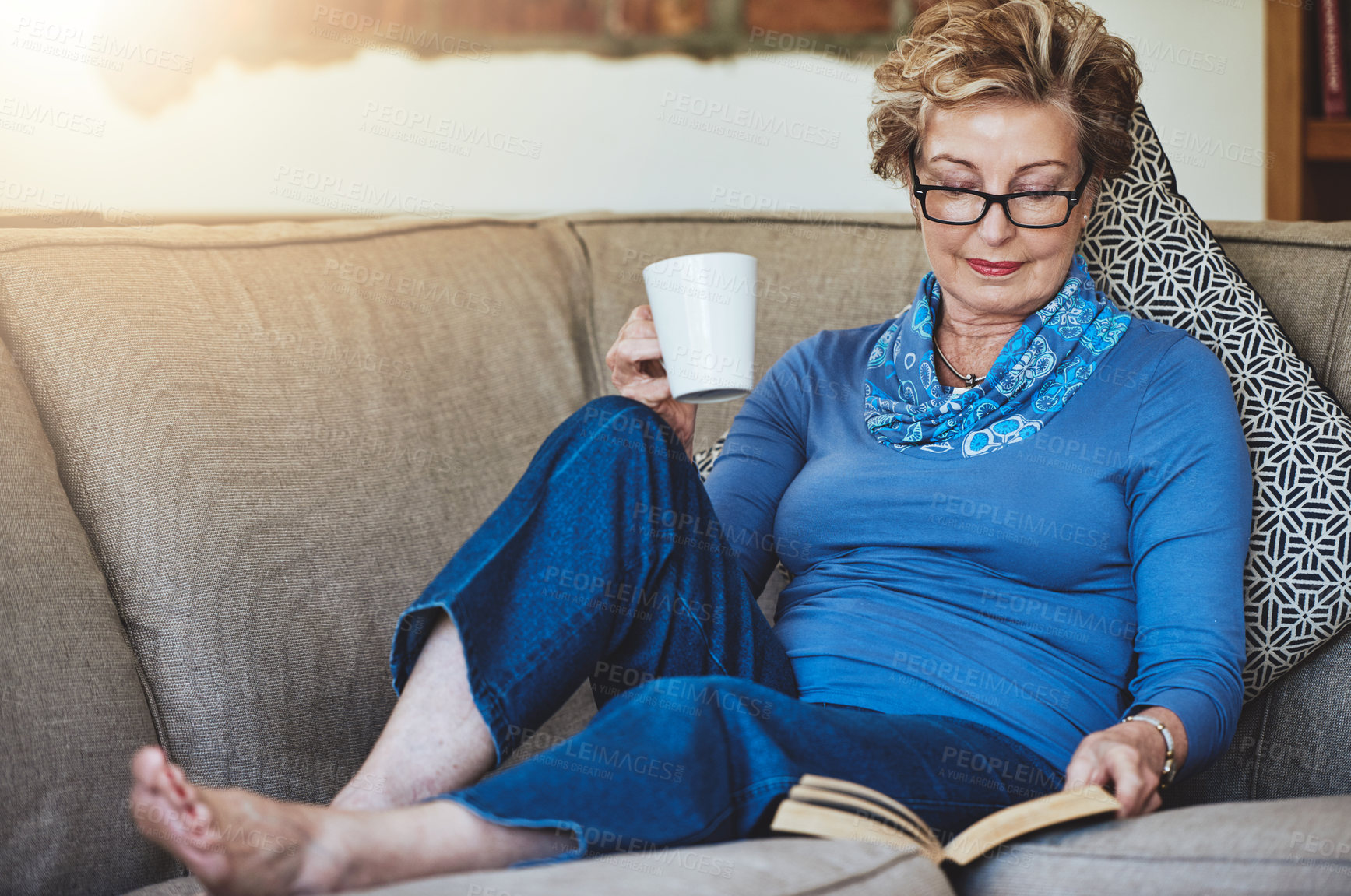 Buy stock photo Shot of a senior woman relaxing with a warm beverage and a book on the sofa at home