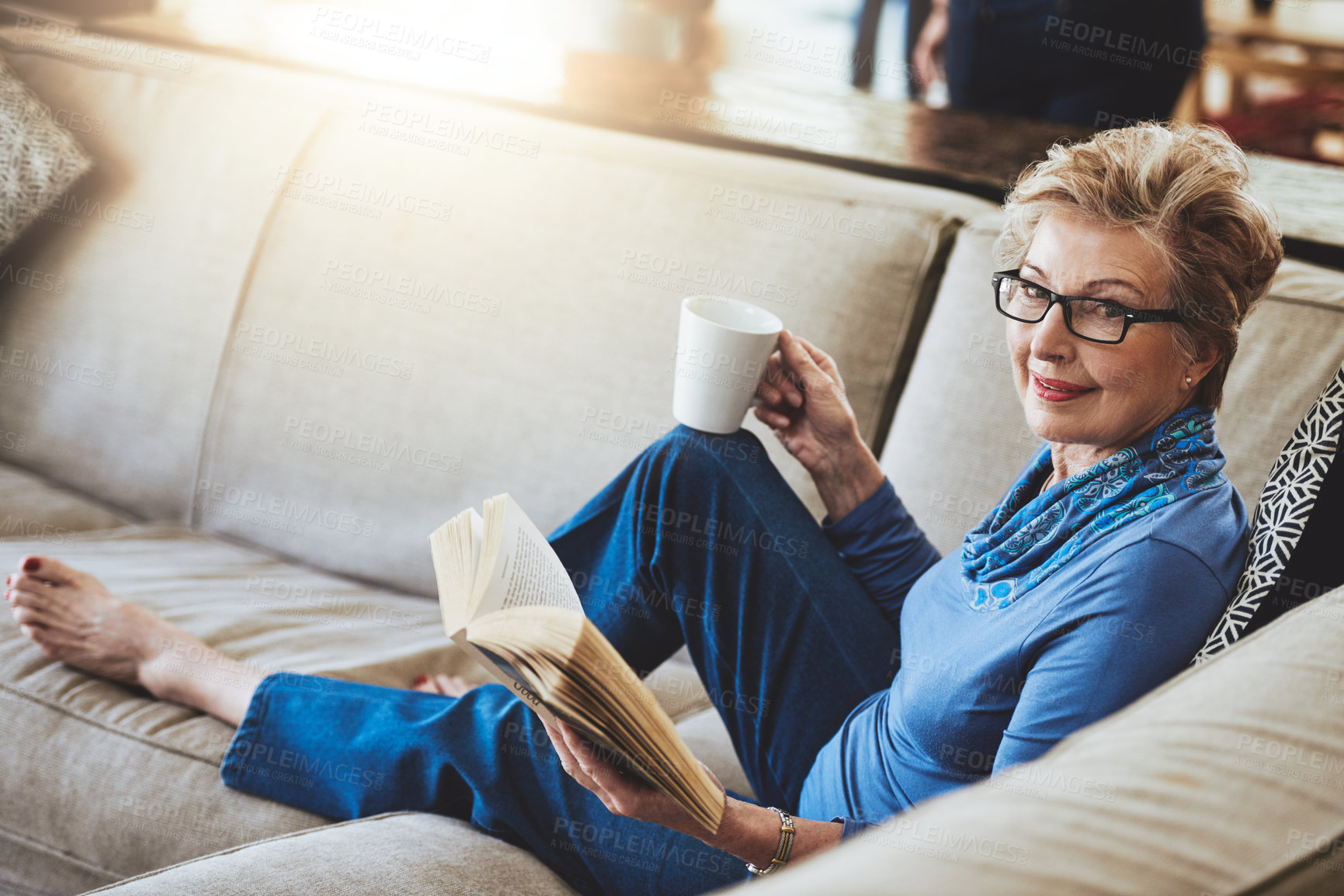 Buy stock photo Portrait of a senior woman relaxing with a warm beverage and a book on the sofa at home