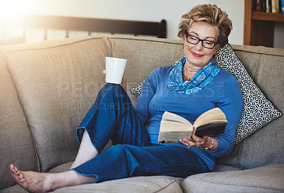 Buy stock photo Shot of a senior woman relaxing with a warm beverage and a book on the sofa at home