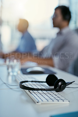 Buy stock photo Closeup shot of a headset lying on a keyboard in an office with call centre agents in the background