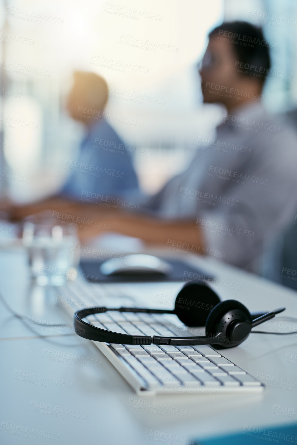 Buy stock photo Closeup shot of a headset lying on a keyboard in an office with call centre agents in the background