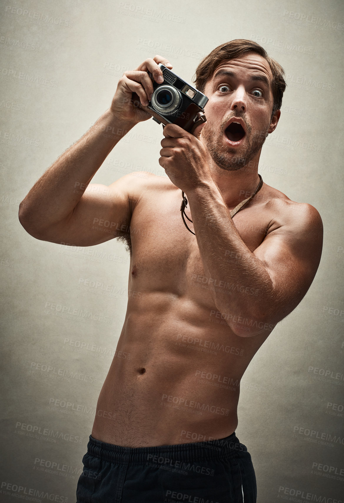 Buy stock photo Studio shot of a shirtless young man taking photos with a vintage camera and looking amazed