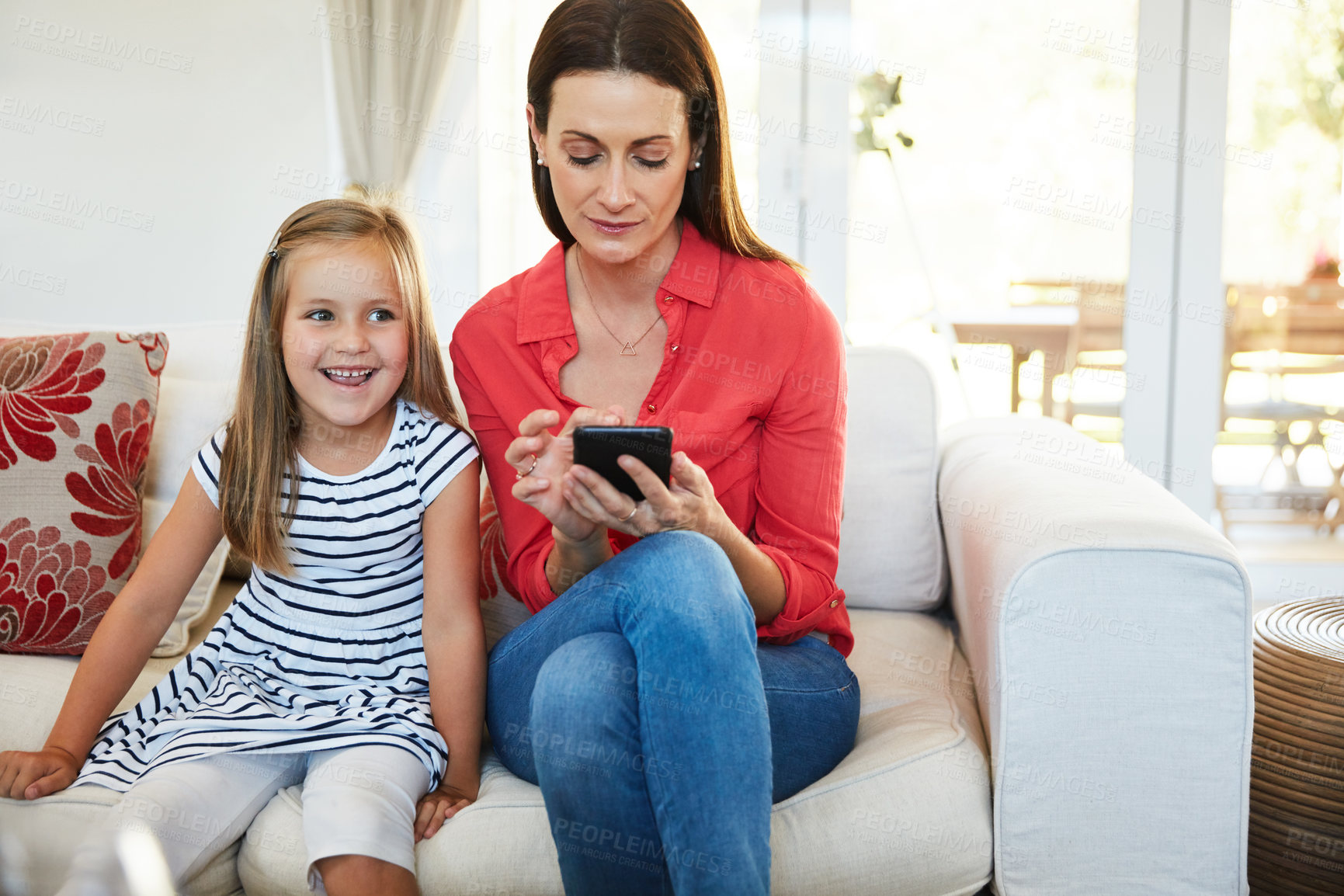Buy stock photo Shot of a mother using a cellphone and her young daughter sitting together in the living room at home