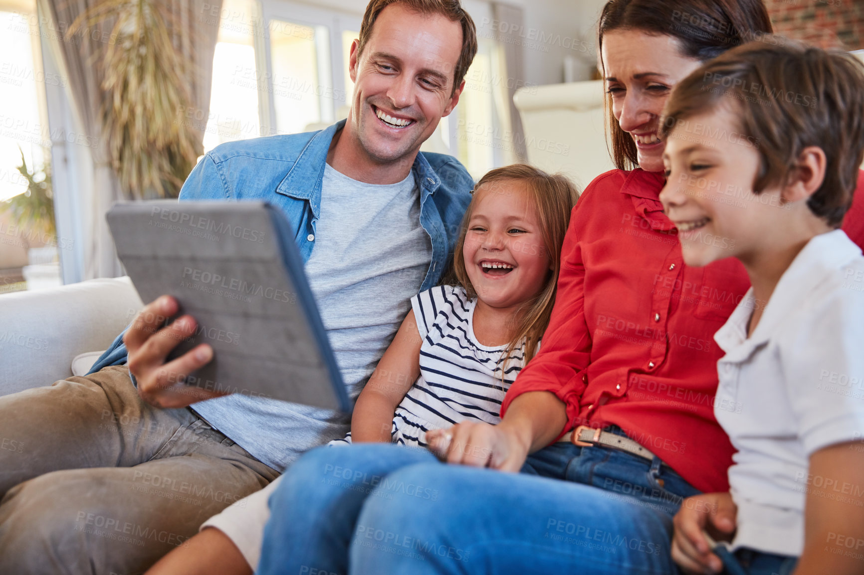 Buy stock photo Shot of smiling parents sitting with their young son and daughter on their living room sofa at home using a digital tablet