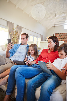Buy stock photo Shot of a young family sitting together on their living room sofa distracted by various media and devices